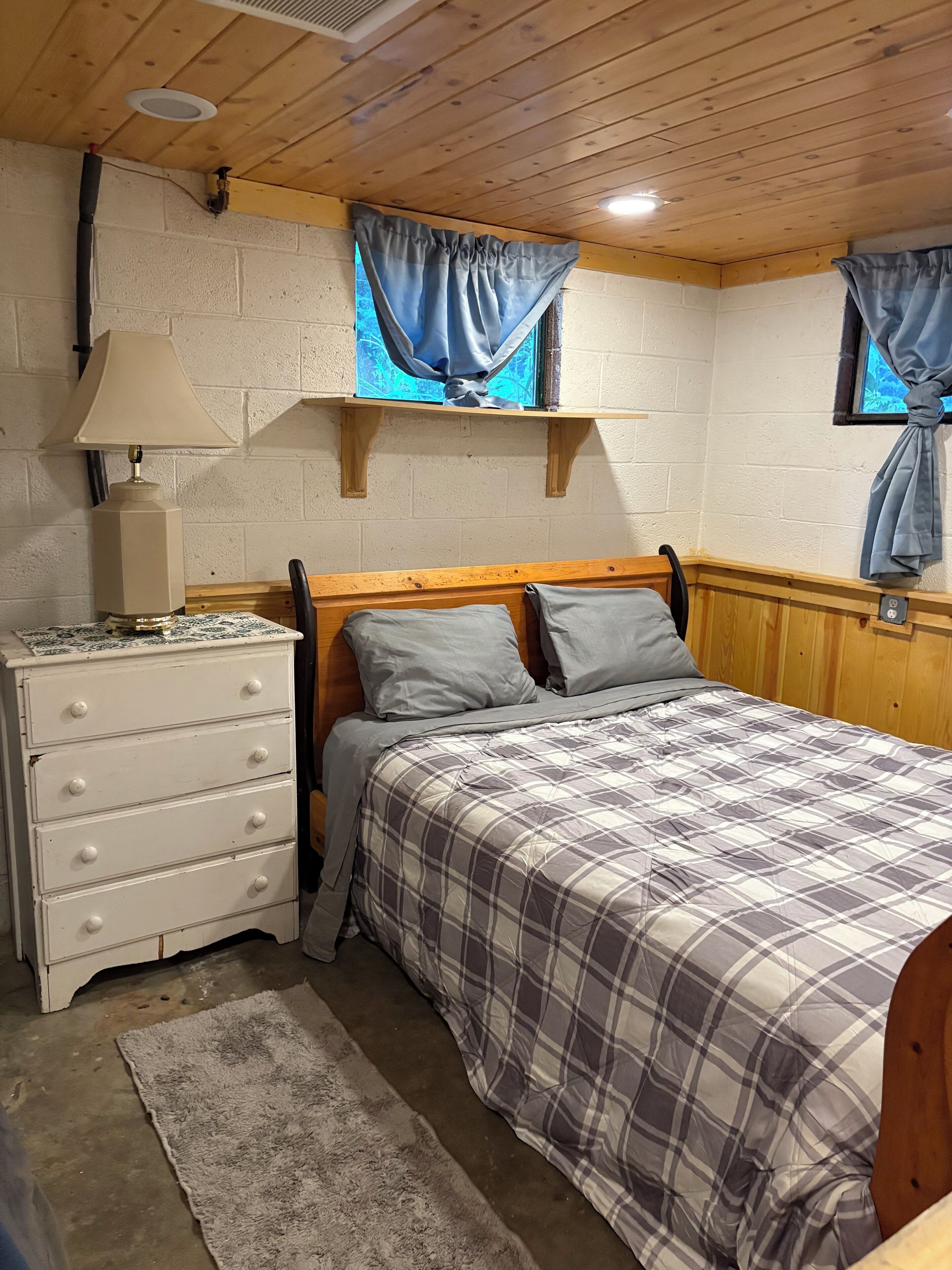 Une chambre à coucher avec une tête de lit en bois, un lit recouvert d’une couverture à carreaux, une commode blanche avec des tiroirs, une lampe et une fenêtre avec des rideaux.