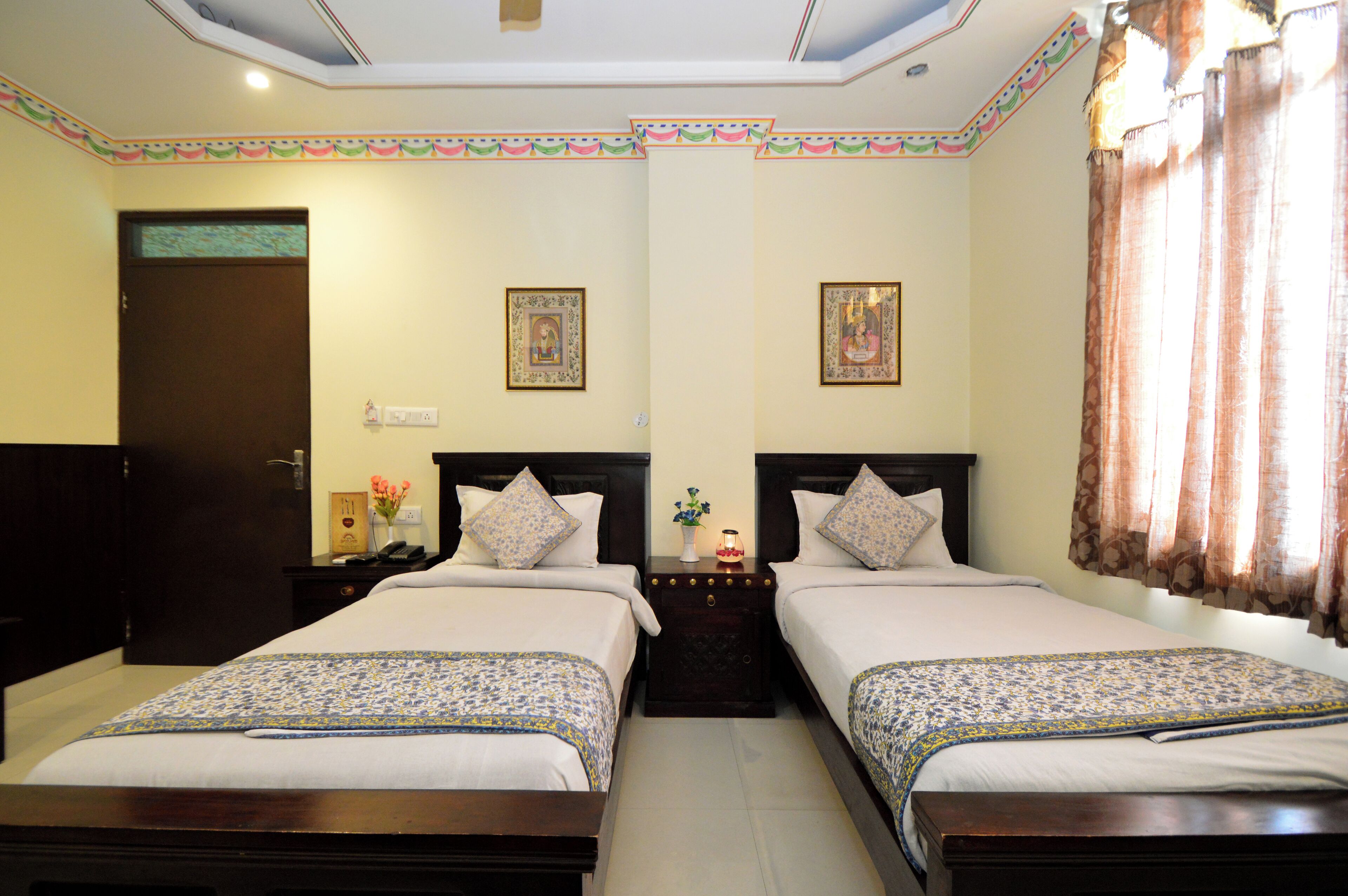 A hotel room with two beds, a wooden headboard, a nightstand, a small table with a vase, and a door.