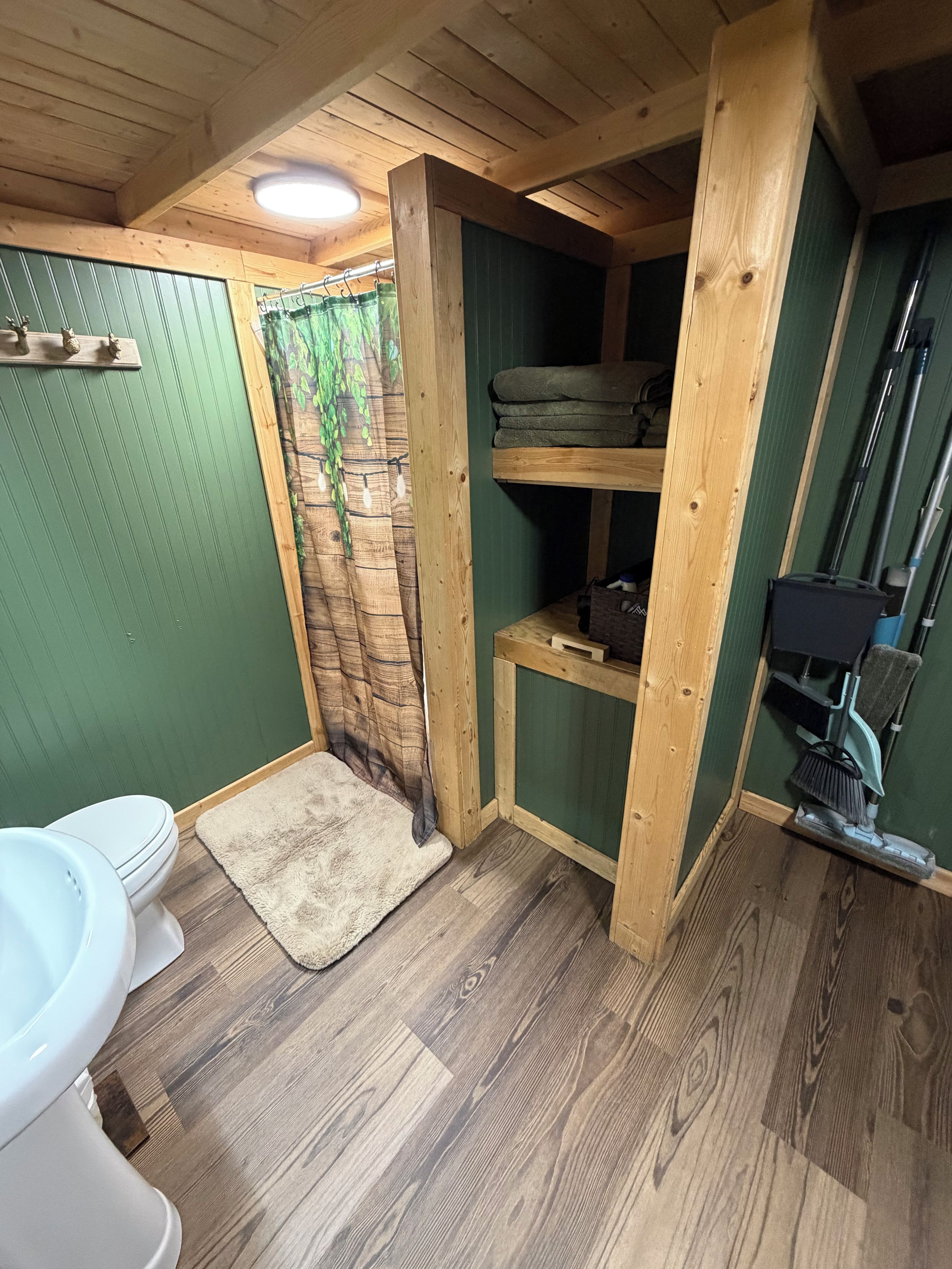 A small, compact bathroom with a wooden ceiling, a green wall, a white toilet, a shower curtain with a leaf pattern, and a wooden shelf with towels.