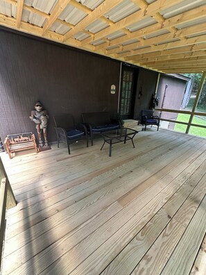 Een overdekt terras buiten met houten vloer, een bank, een tafel en een beeld.