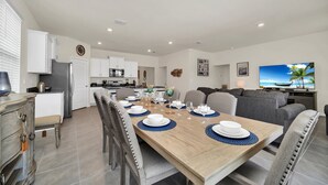 A modern kitchen with white cabinets, a refrigerator, and a microwave. There is a dining area with a wooden table set for four, surrounded by chairs. A gray sofa and a television are visible in the background. - Magical Family Getaway 7BD Villa with (Davenport)