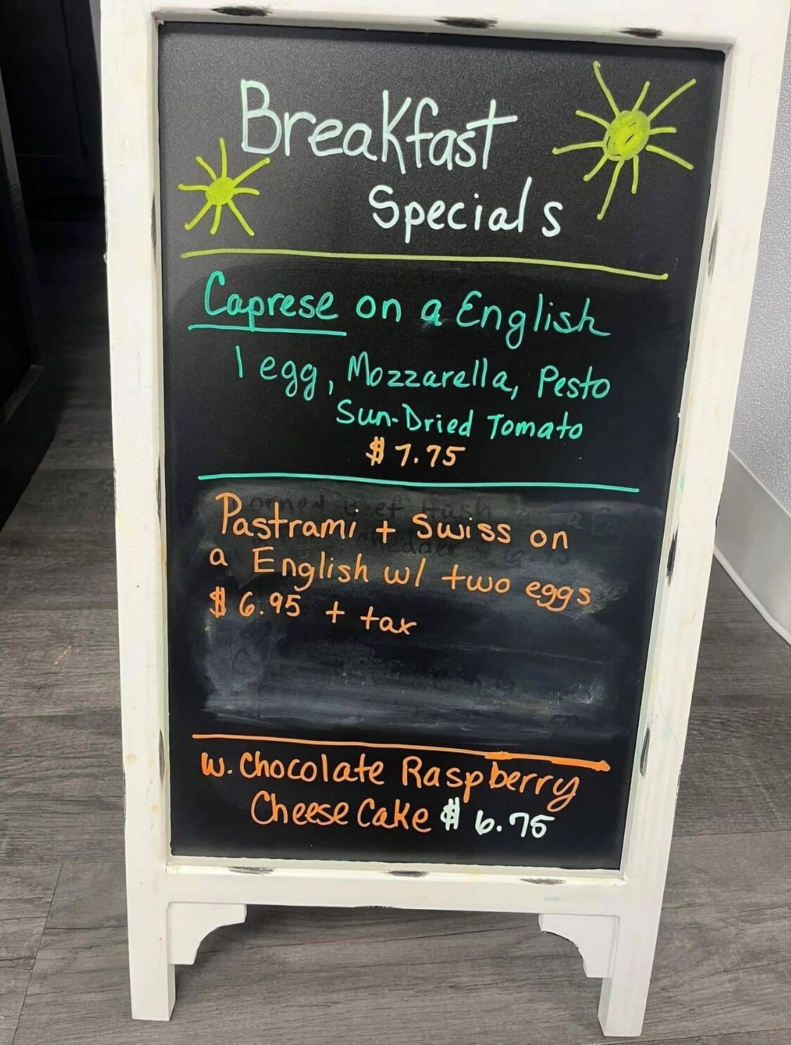 Un tableau noir affiche le menu du petit-déjeuner, avec des plats spéciaux comme le « Caprese on English », le « Pastrami et fromage suisse » et le « gâteau au fromage au chocolat et aux framboises ».