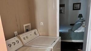 A laundry area with a washer and dryer, a small mirror, and a bed in the adjacent room. - Home Away From Home 2 (East Palatka)