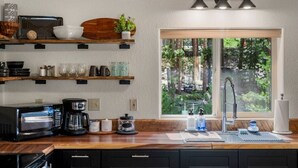 A kitchen with wooden shelves, a coffee maker, a microwave, and a window with a view of trees.