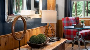 A cozy room with a wooden table, a bowl of greenery, a lamp, a book titled 'Martha Stewart's Cooking School', a plaid sofa, and a mirror with an ornate frame.
