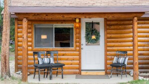 A log cabin with a front porch, two chairs, a door with a wreath, and a window.