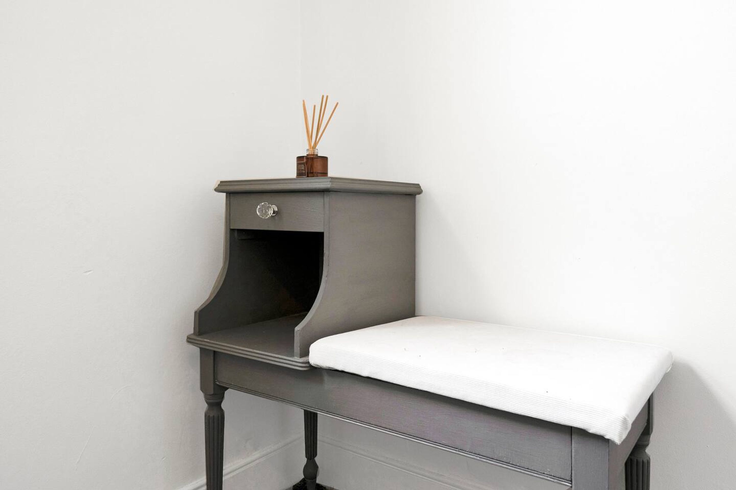 A small, dark-colored nightstand with a drawer, a white cushion, and a small container with reed sticks on top.