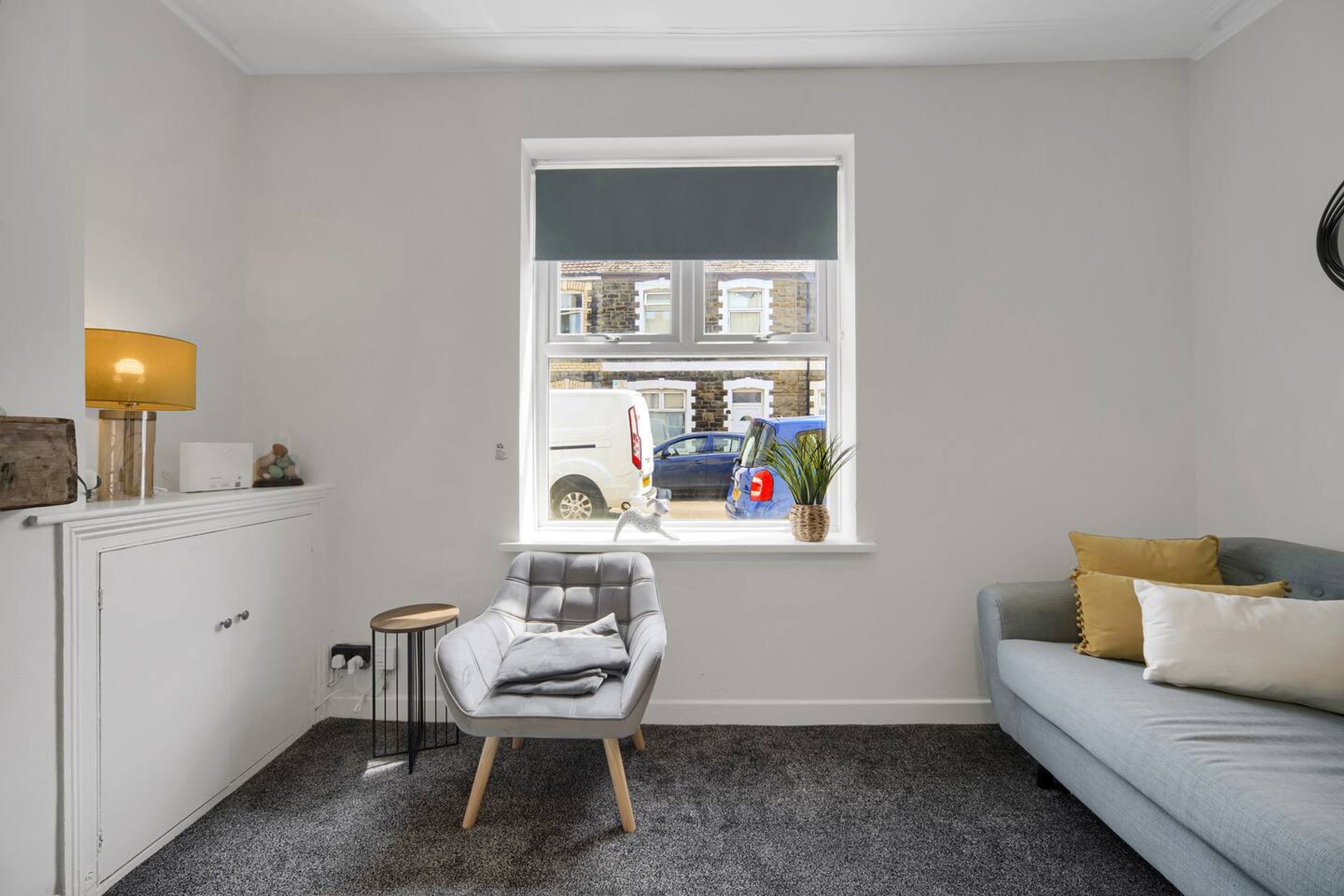 A living room with a grey sofa, a small armchair, a white cabinet, a lamp, and a window with a view of a street.