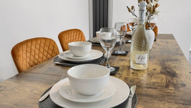 Een eettafel gedekt met witte kommen, borden, bestek en een flesje Prosecco.