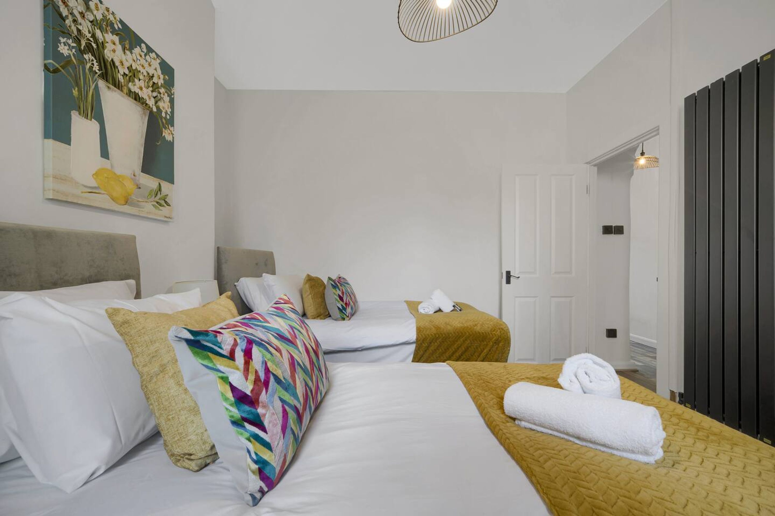 Une chambre d’hôtel avec deux lits, du linge de lit blanc, un plaid coloré, un tableau au mur et une porte sur laquelle est posée une serviette.