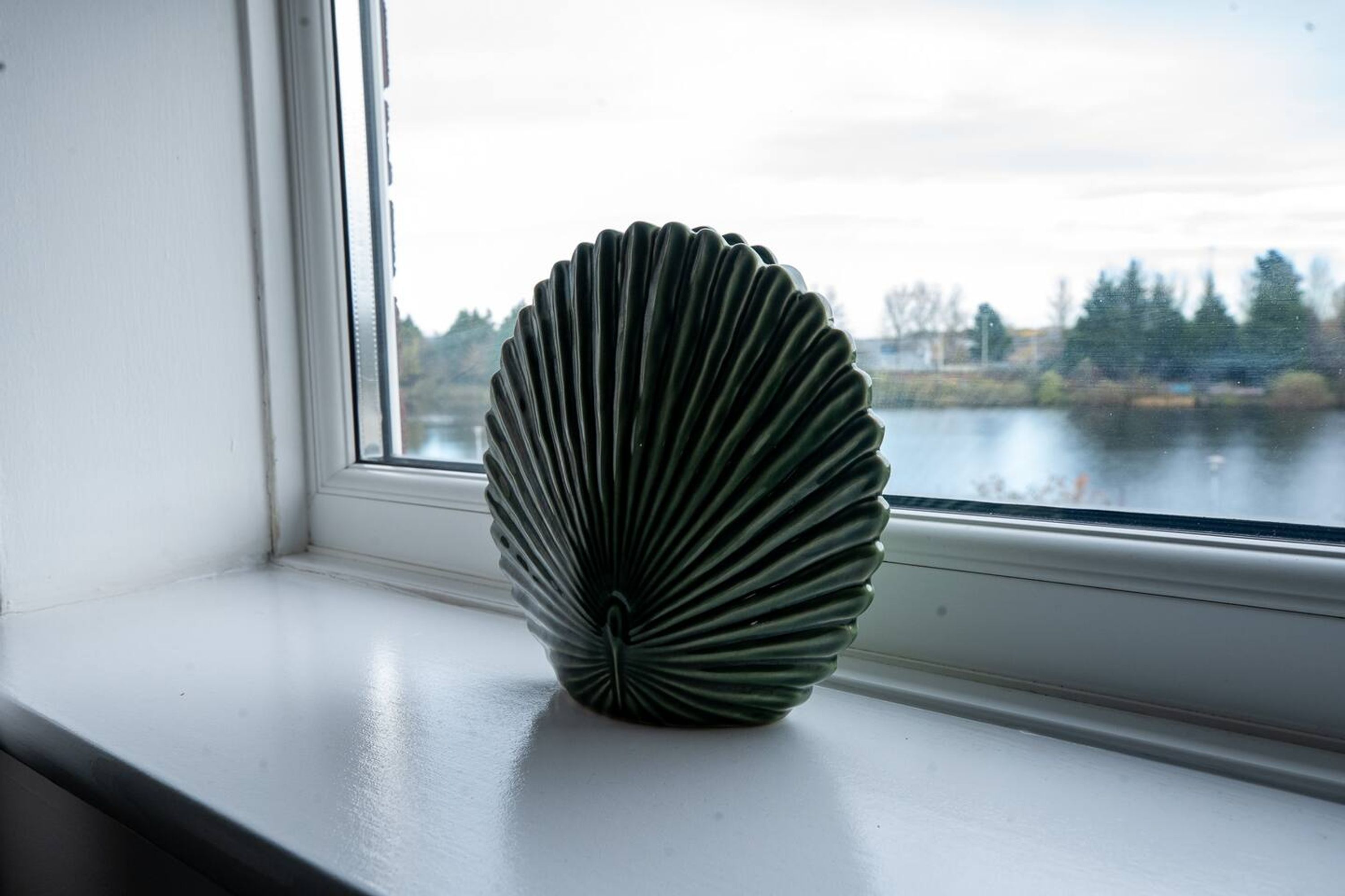 Un vase en forme de coquillage, posé sur un rebord de fenêtre, d’où l’on aperçoit un lac et des arbres.
