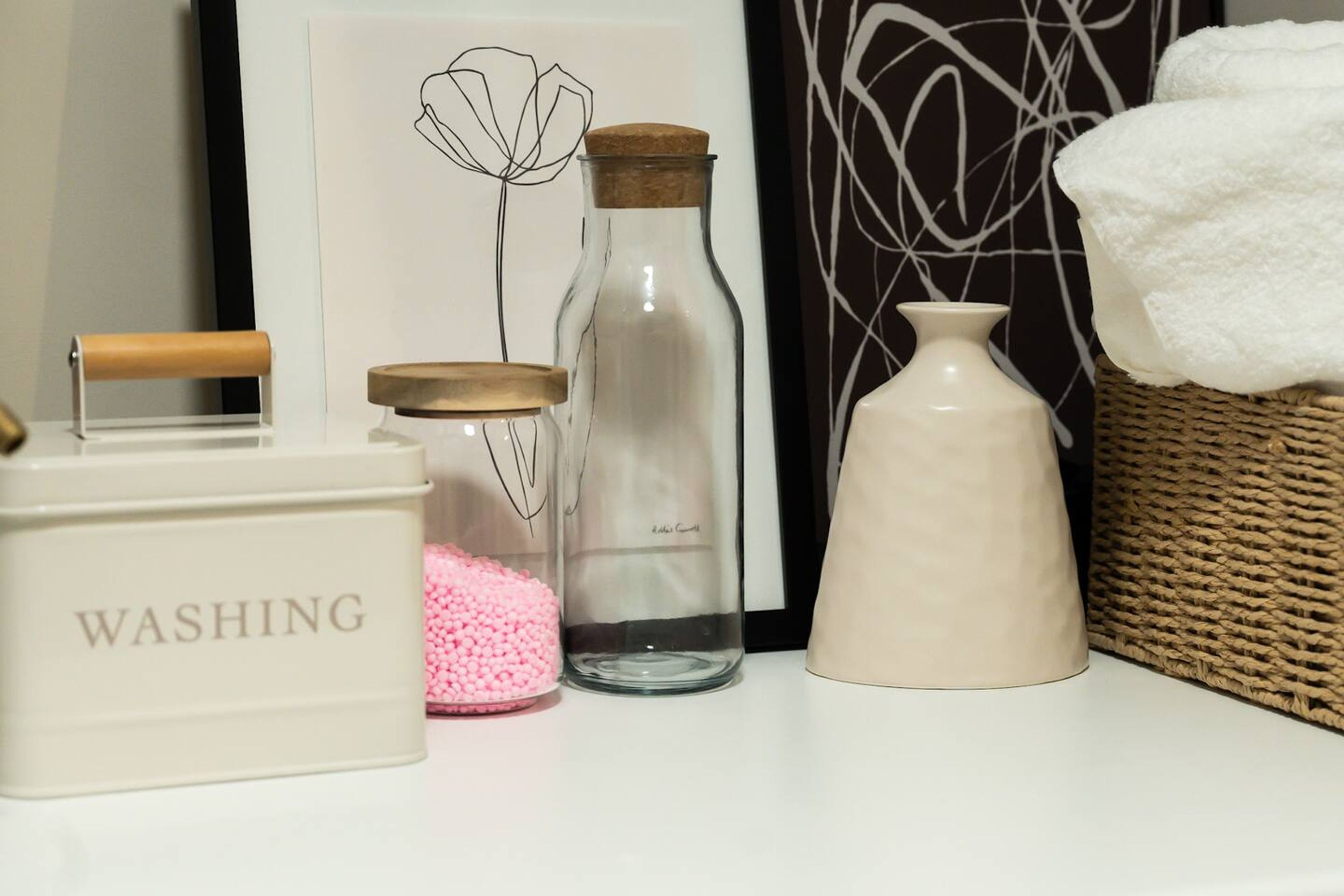 A bathroom vanity with a "WASHING" container, a glass bottle with a cork, a clear glass jar with pink beads, and a white ceramic container.