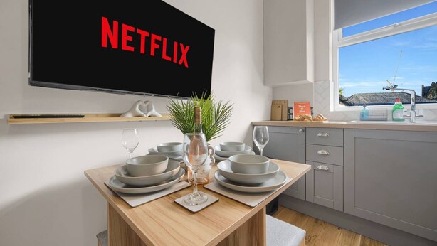 A modern kitchen with grey cabinets, a wooden dining table set with plates, bowls, and a vase with a plant. A large TV screen displays the Netflix logo. - Free Parking Fast Wifi 2 Min to Cardiff City (Cardiff)