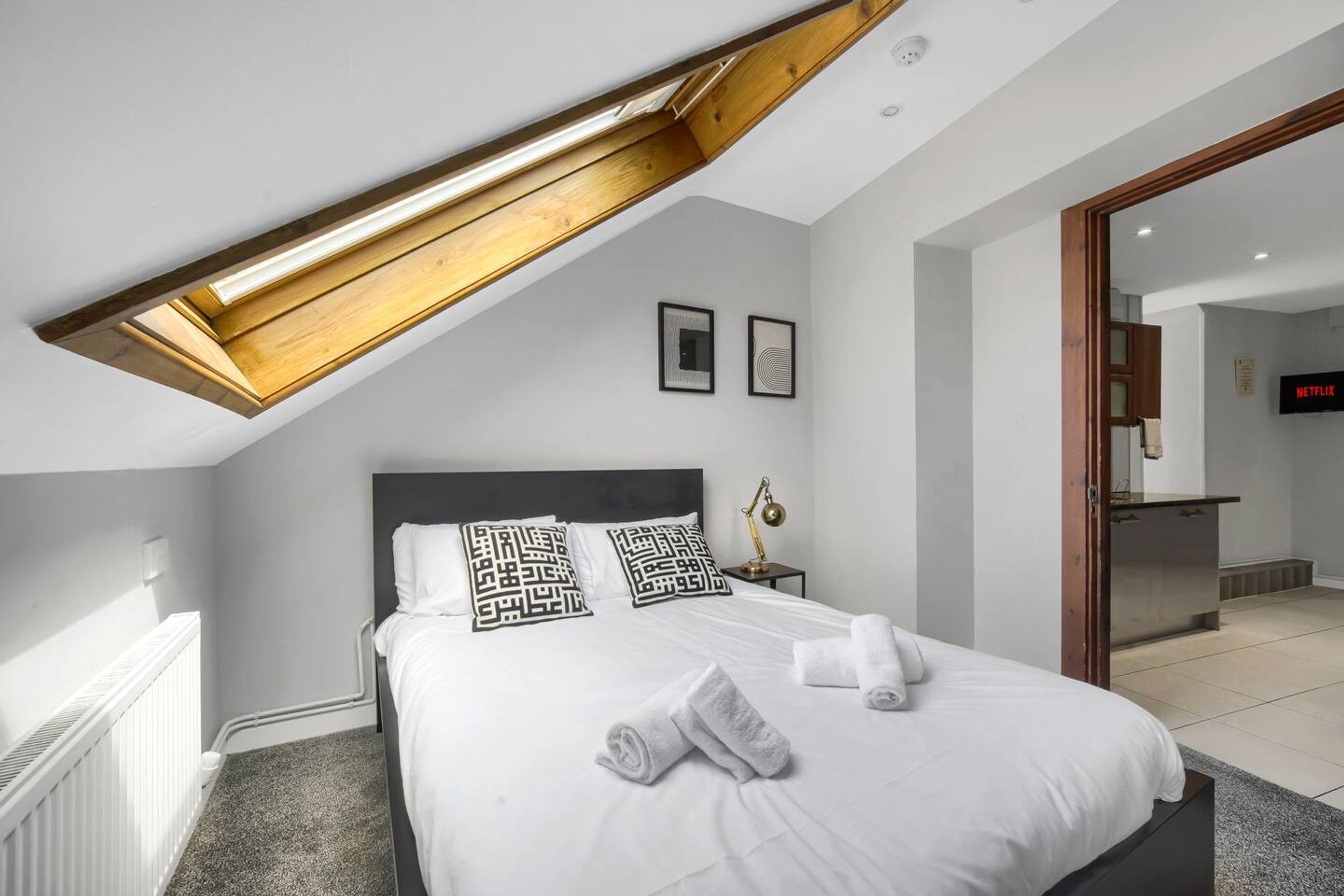 A modern bedroom with a skylight, a bed with decorative pillows, a bedside lamp, and a view of the kitchen area.