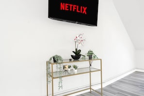 A modern living room with a glass console table, a black TV mounted on the wall, and a variety of potted plants. - Luxury Penthouse Suite - Swansea Central - Beach (Swansea)