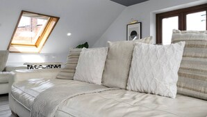 Un dormitorio tipo loft moderno con techo inclinado, un ventanal grande, un sofá blanco y una cama con muchas almohadas.