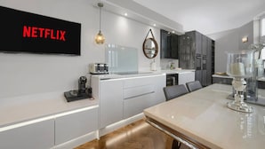 A modern kitchen with a large flat-screen TV displaying "NETFLIX," a built-in oven, and a dining area with a marble table. - Modern Unique - Central Beach Access (Swansea)