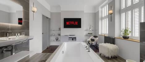 Un baño moderno con una bañera grande, un tocador con espejo y un televisor de pared mostrando "NETFLIX".