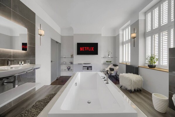 A modern bathroom with a large bathtub, a vanity with a mirror, and a wall-mounted television displaying "NETFLIX". - Modern Unique - Central Beach Access (Swansea)