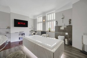 A modern bathroom with a large bathtub, a toilet, and a sink. There is a TV mounted on the wall displaying 'NETFLIX'. - Modern Unique - Central Beach Access (Swansea)