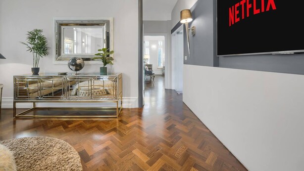A modern living room with a herringbone wood floor, a glass coffee table, a mirror, and a flat-screen TV mounted on the wall. - Modern Unique - Central Beach Access (Swansea)