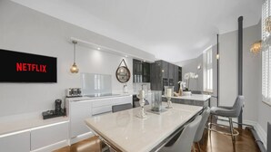 A modern kitchen with a large island, white cabinetry, and a flat-screen TV displaying 'NETFLIX'. - Modern Unique - Central Beach Access (Swansea)