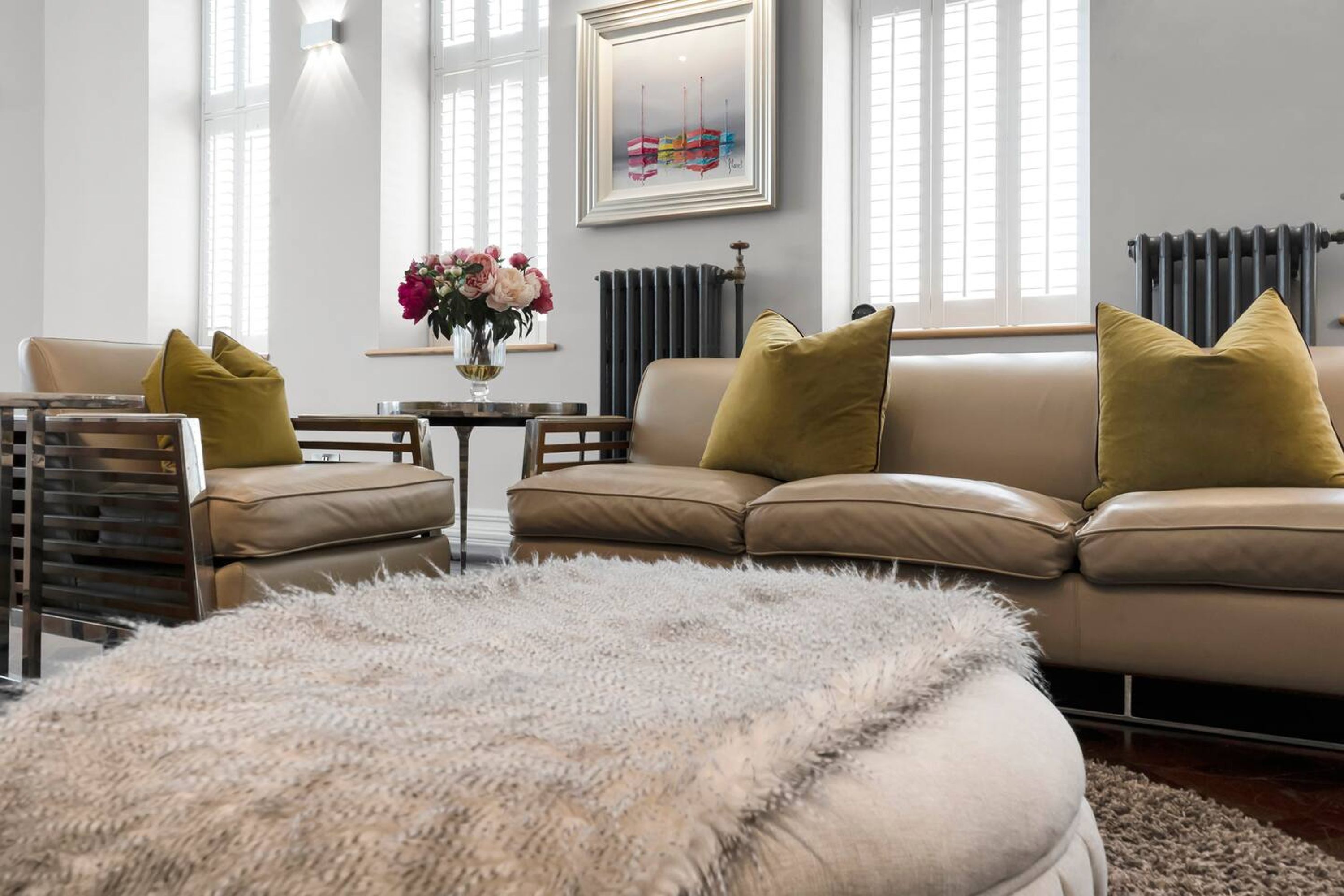 A living room with a beige sofa, two yellow cushions, a fluffy white rug, a wooden side table with a vase of flowers, and a framed artwork on the wall.
