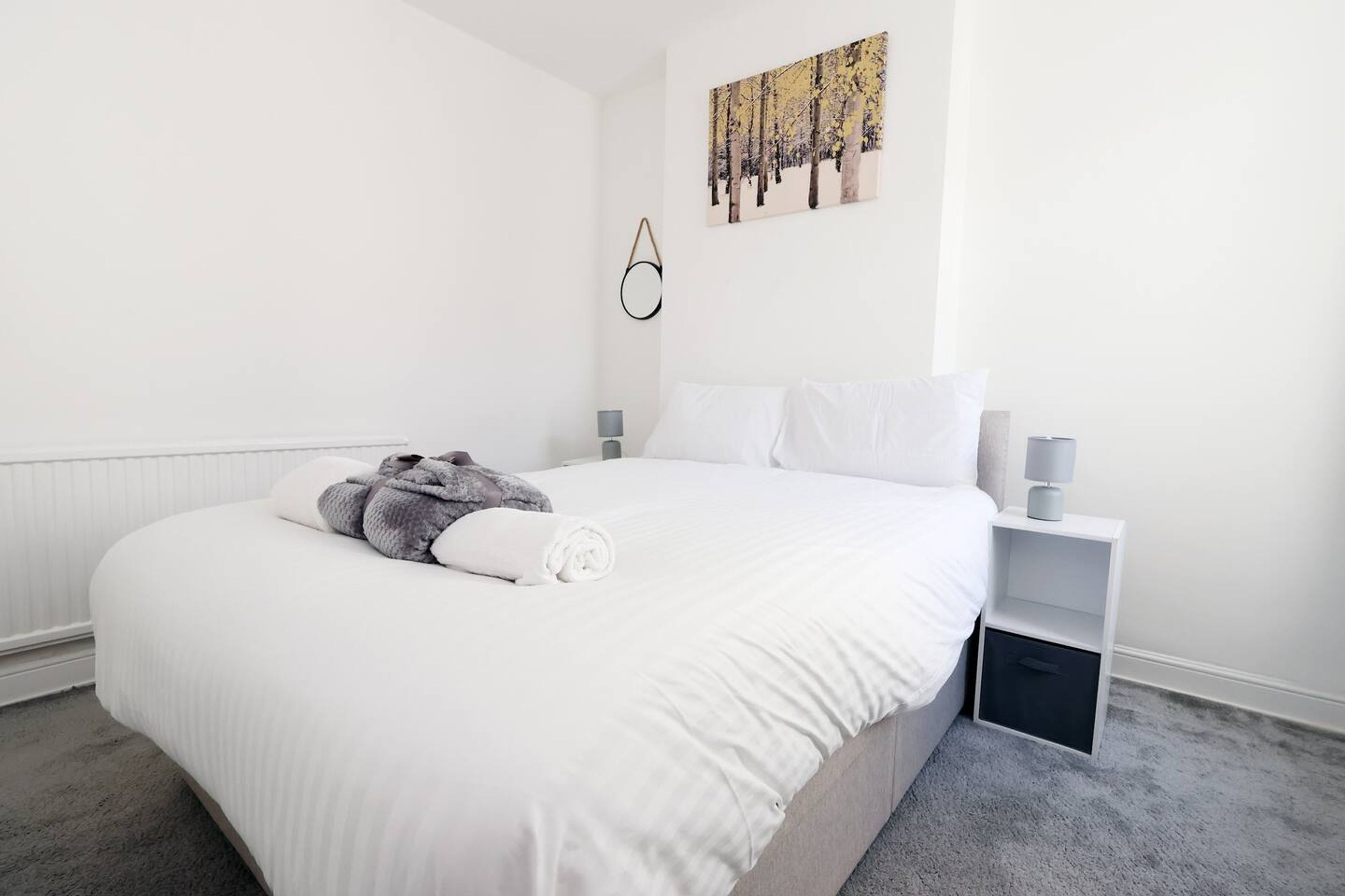 Ein ordentlich bezogenes Bett mit weißen Laken und einem zusammengerollten Handtuch, ein Nachttisch mit Lampe sowie ein Wandbild.