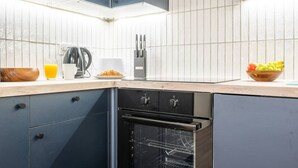 A modern kitchen with dark cabinetry, a built-in oven, and a countertop with a kettle, coffee maker, and fruit bowl. - Luxurious 2 Bed Central Cardiff Free EV Charge (Cardiff)