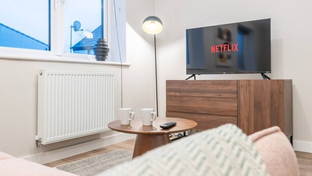 A modern living room with a television displaying 'NETFLIX', a round wooden table with two white mugs, a remote control, and a radiator. - Luxurious 2 Bed Central Cardiff Free EV Charge (Cardiff)