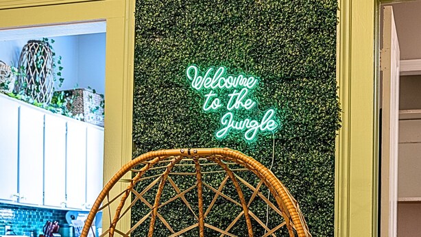 A wicker chair with a cushion in a room with a green wall and a neon sign that reads "Welcome to the Jungle." - The Jungle Nook: Themed,Steps to Forsyth,King Bed,Grill,Games,Picnic Basket (Savannah)