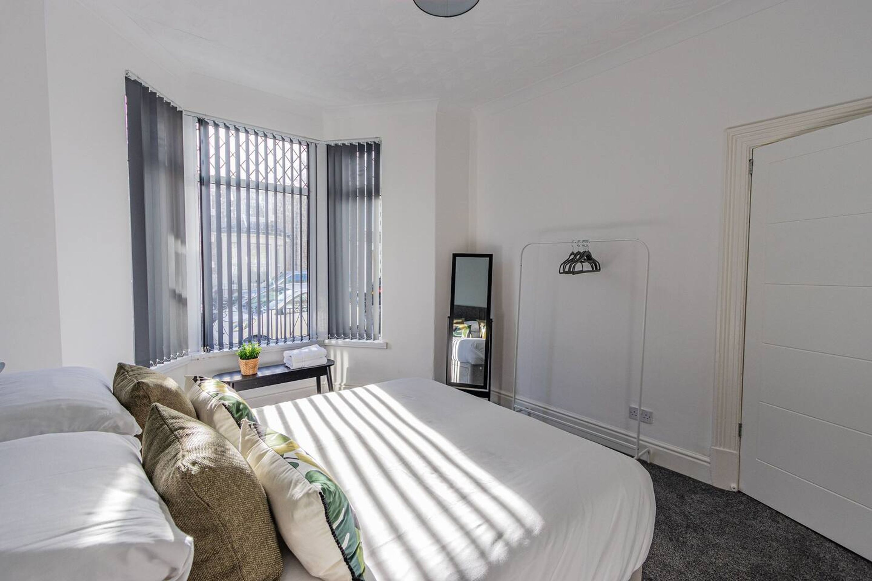 A bedroom with a large bed, a window with blinds, a small table with a plant, and a mirror on the wall.