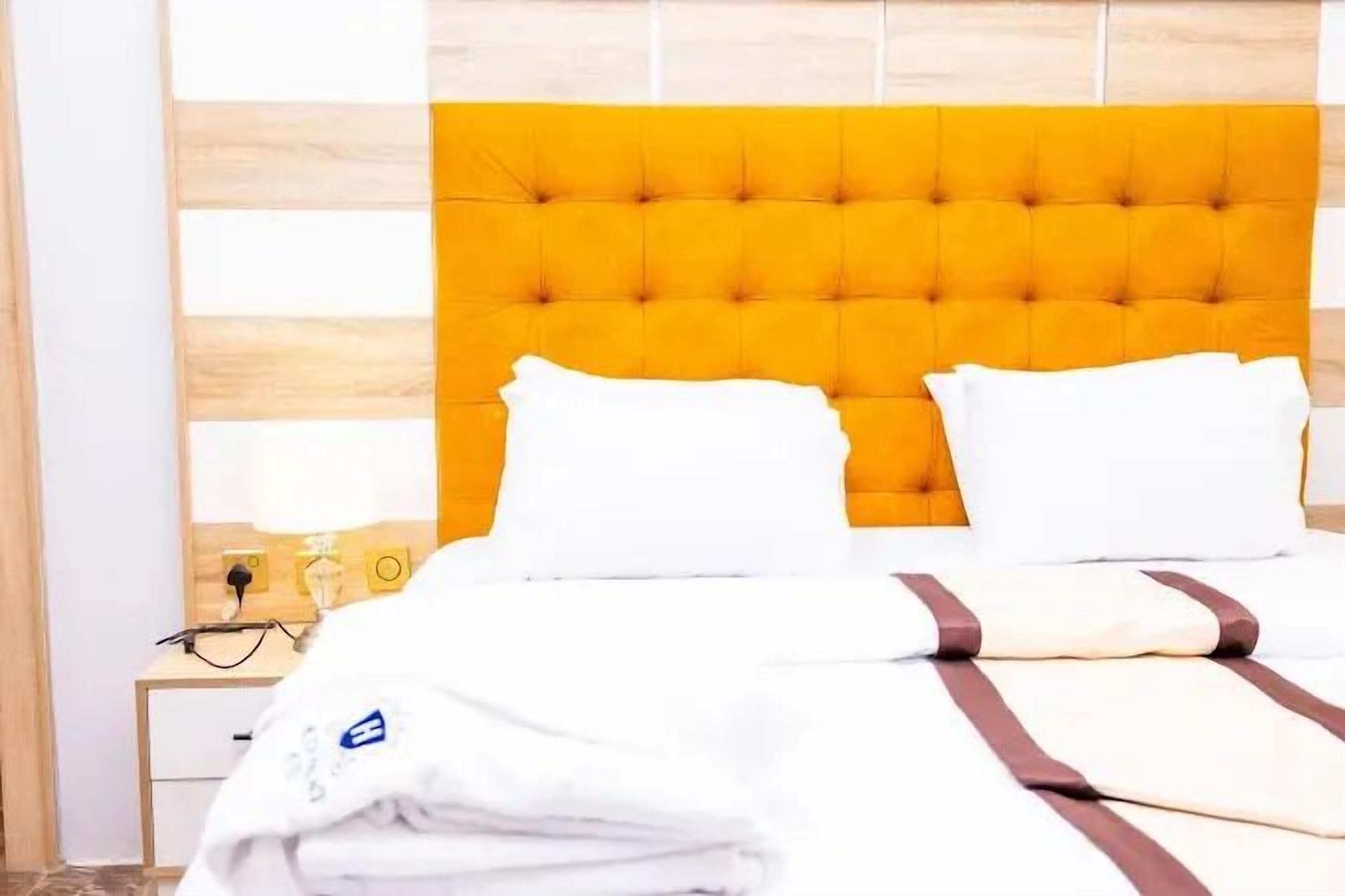 Ein Hotelzimmer mit einem gesteppten, gelben Kopfteil, weißer Bettwäsche und einem Nachttisch aus Holz mit Lampe und Telefon.