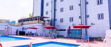 A hotel with a swimming pool, red umbrellas, and a multi-story building.