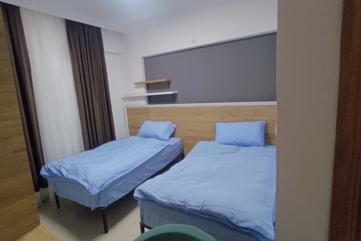 A room with two single beds, a wooden wardrobe, a small shelf, and a curtain.