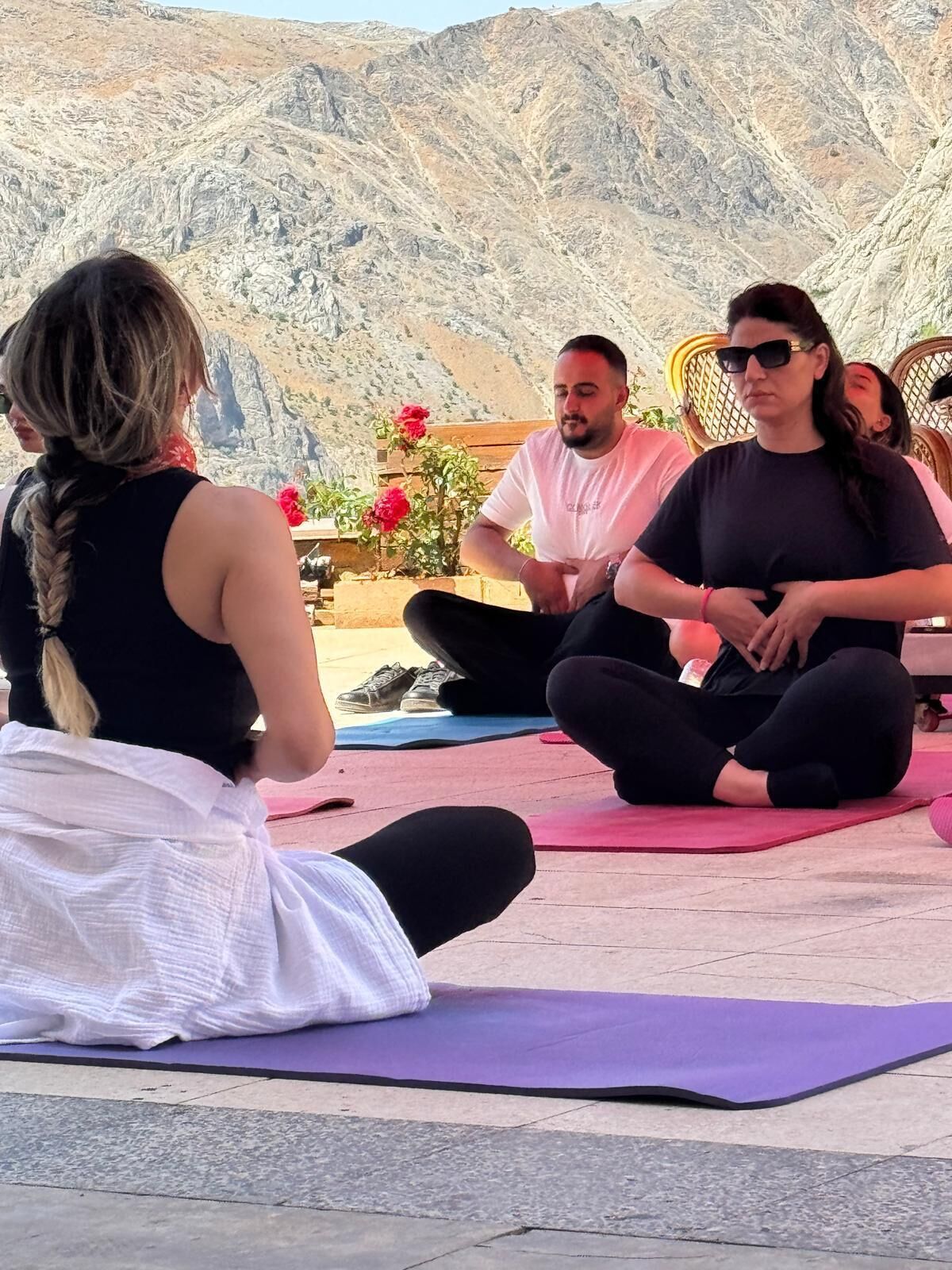A group of people sitting on yoga mats outdoors with a mountainous backdrop.