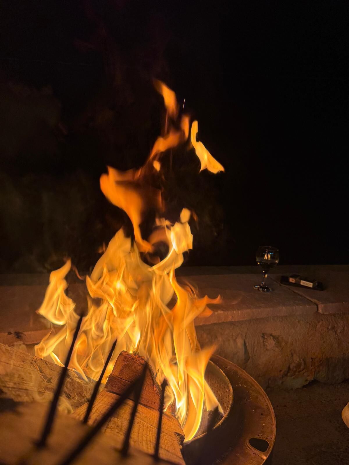A campfire with logs and burning flames, a glass of wine, and a small box on a concrete surface at night.