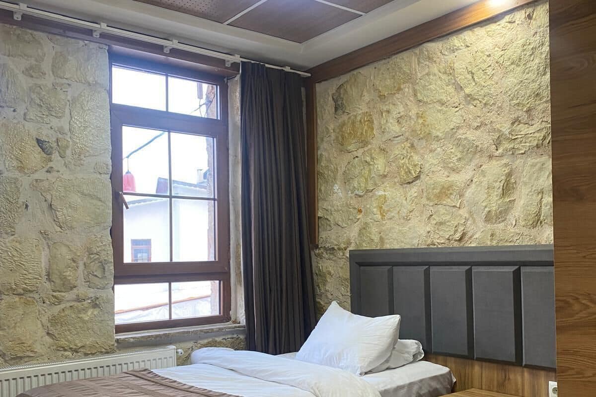 A bedroom with a stone wall, a bed with a striped blanket, a bedside table, and a window with curtains.
