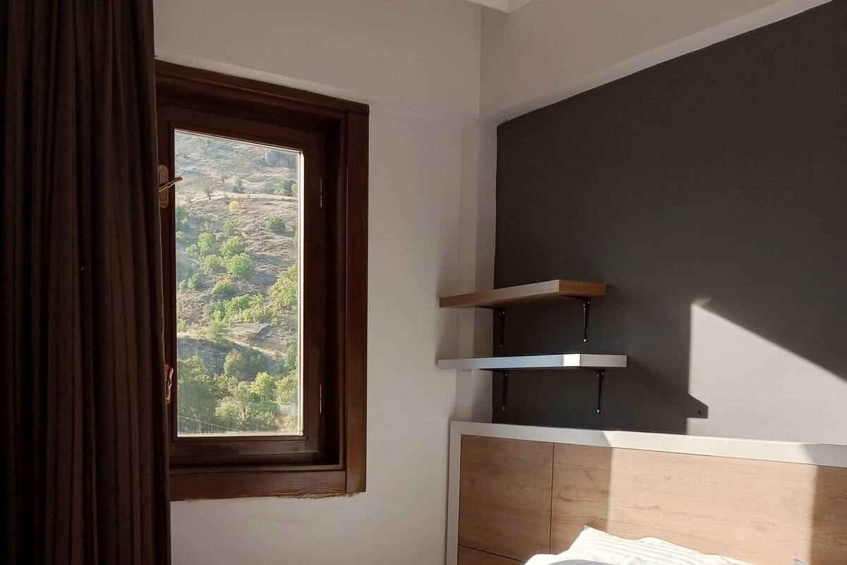 A hotel room with a bed, a window with curtains, a headboard, and a shelf unit.