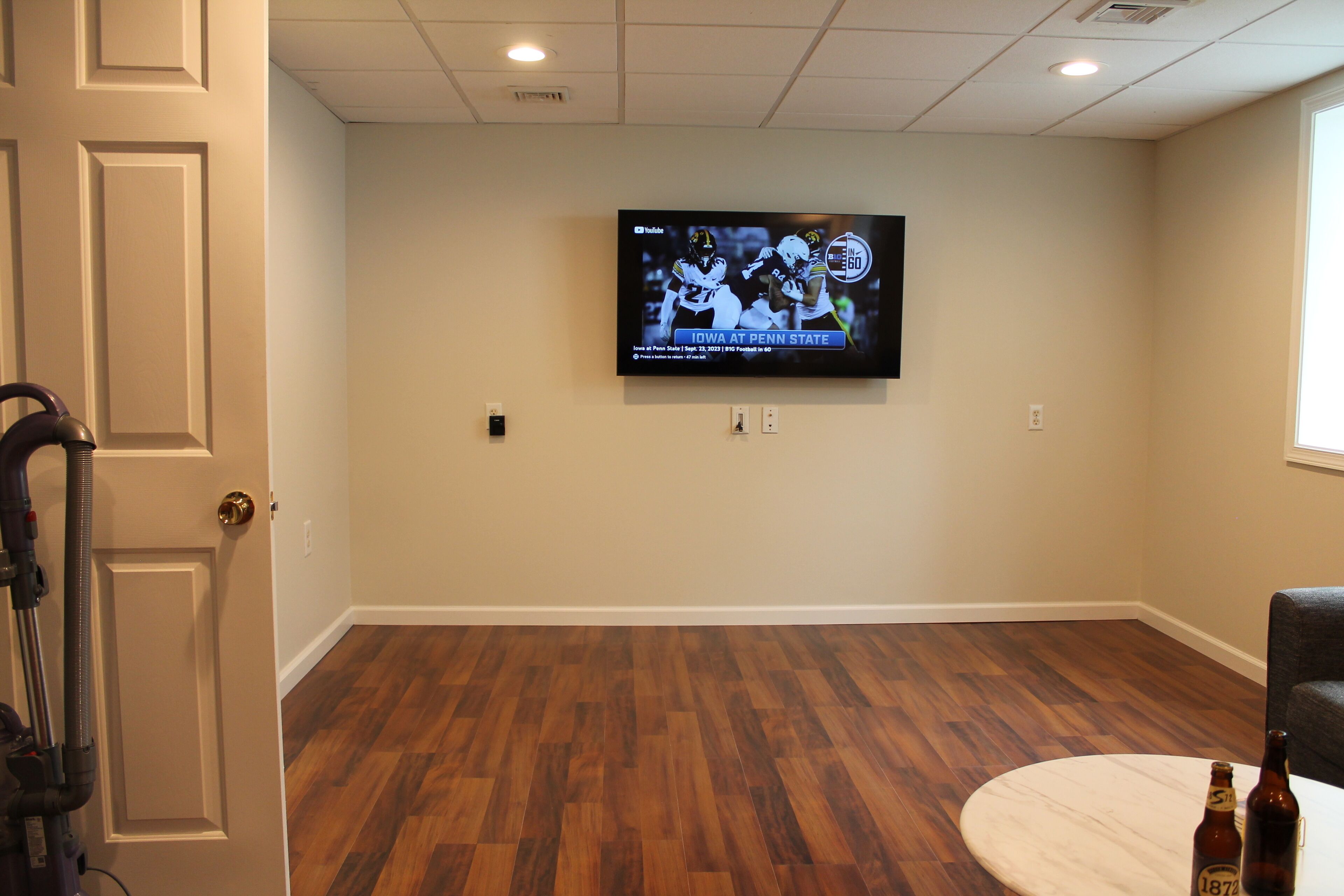 A room with a wooden floor, a television mounted on the wall, and a door with a vacuum cleaner.