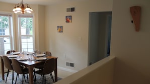 En spiseplads med et træbord dækket til seks personer, omgivet af stole. To vinduer med persienner, en loftlampe og et vægur.
