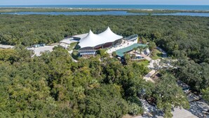 An aerial view of a resort with a distinctive white tent-like structure surrounded by dense forest and a body of water in the background.
