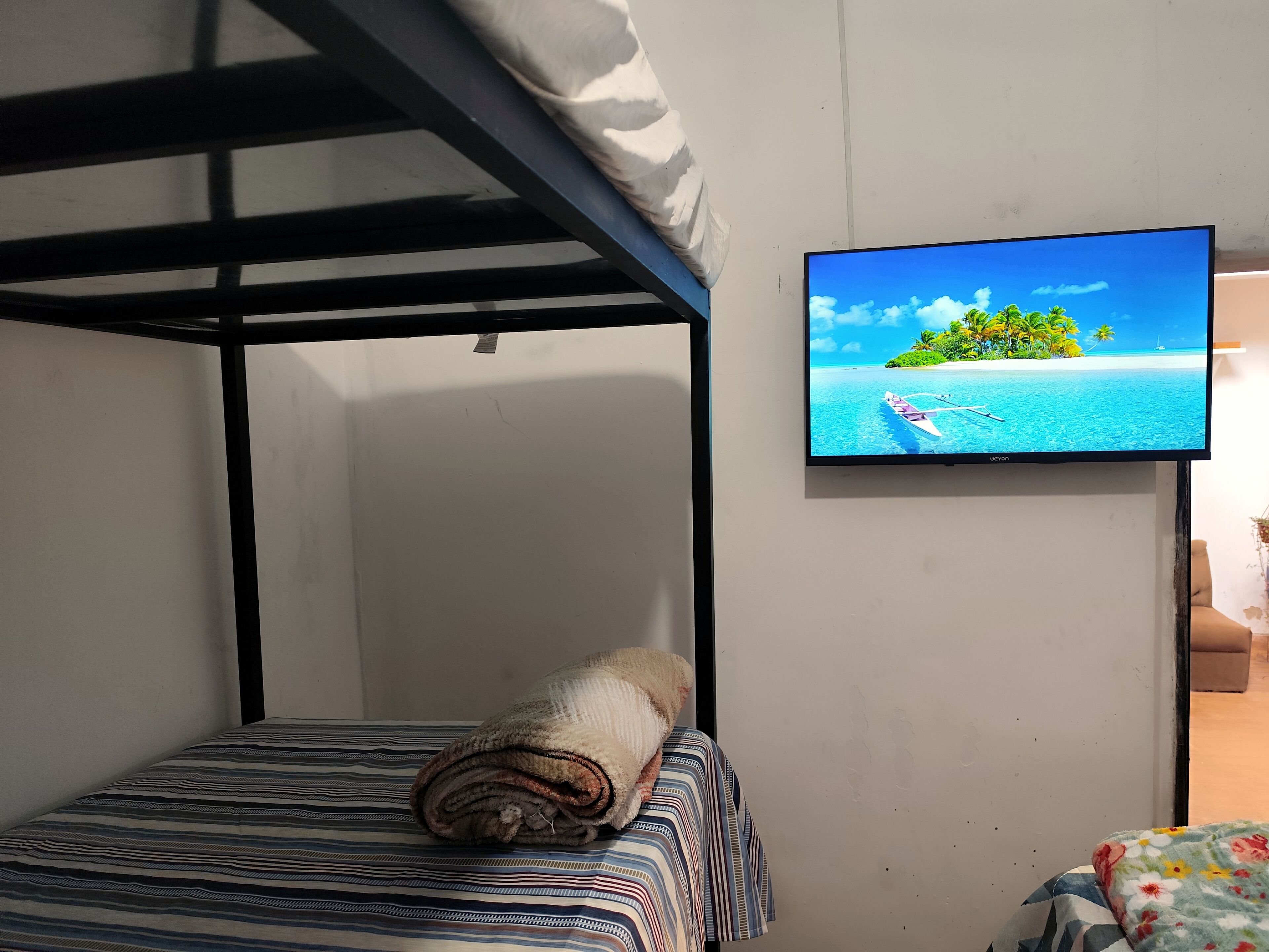 Yhden hengen sänky, jossa on raidallinen päiväpeite, rullalle taitettu pyyhe ja televisio, josta näkyy kuva trooppisesta rannasta.