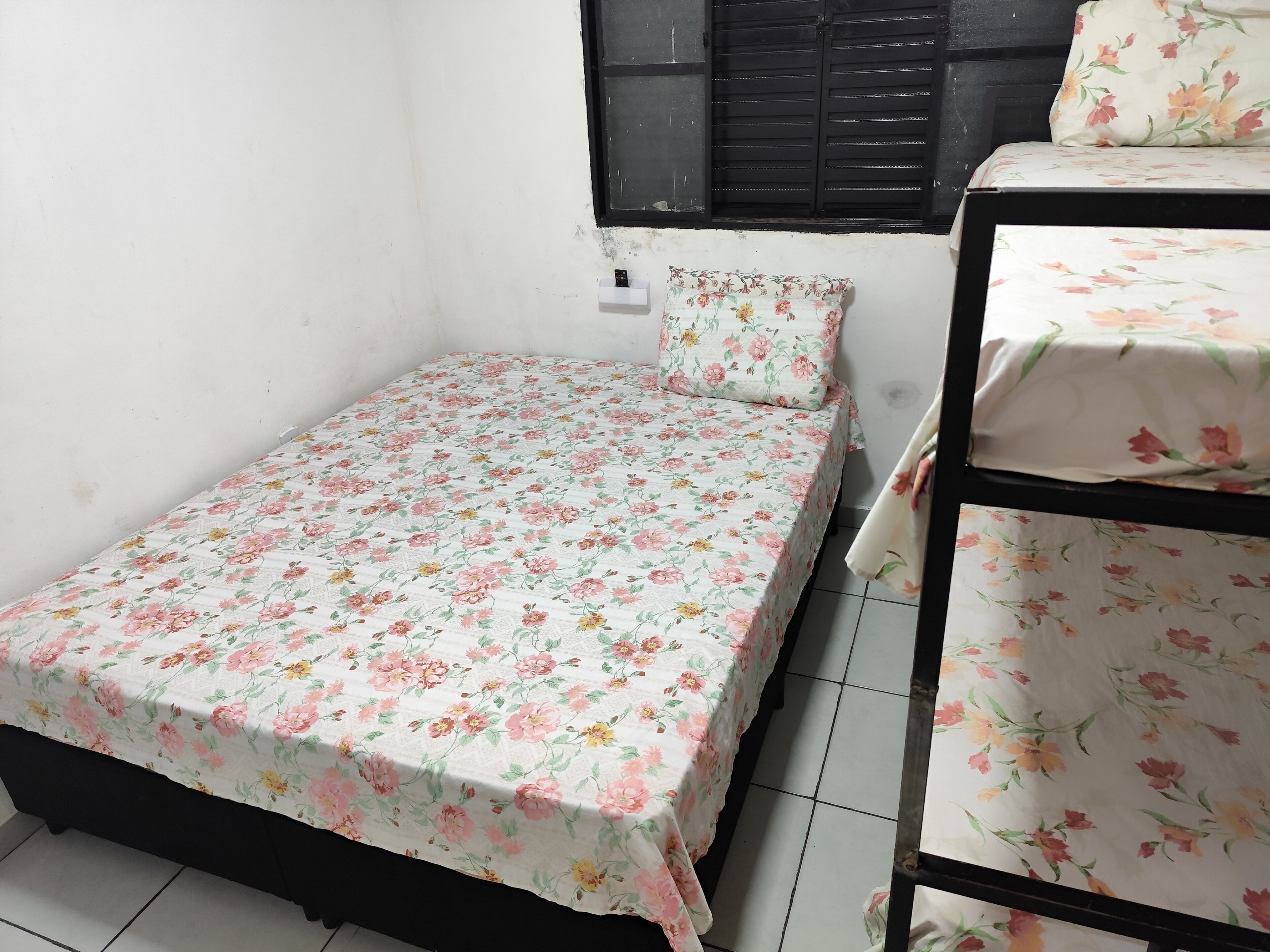 Una habitación pequeña con cama individual, edredón estampado con motivos florales, estructura de cama de metal negro y ventana con contraventanas negras.
