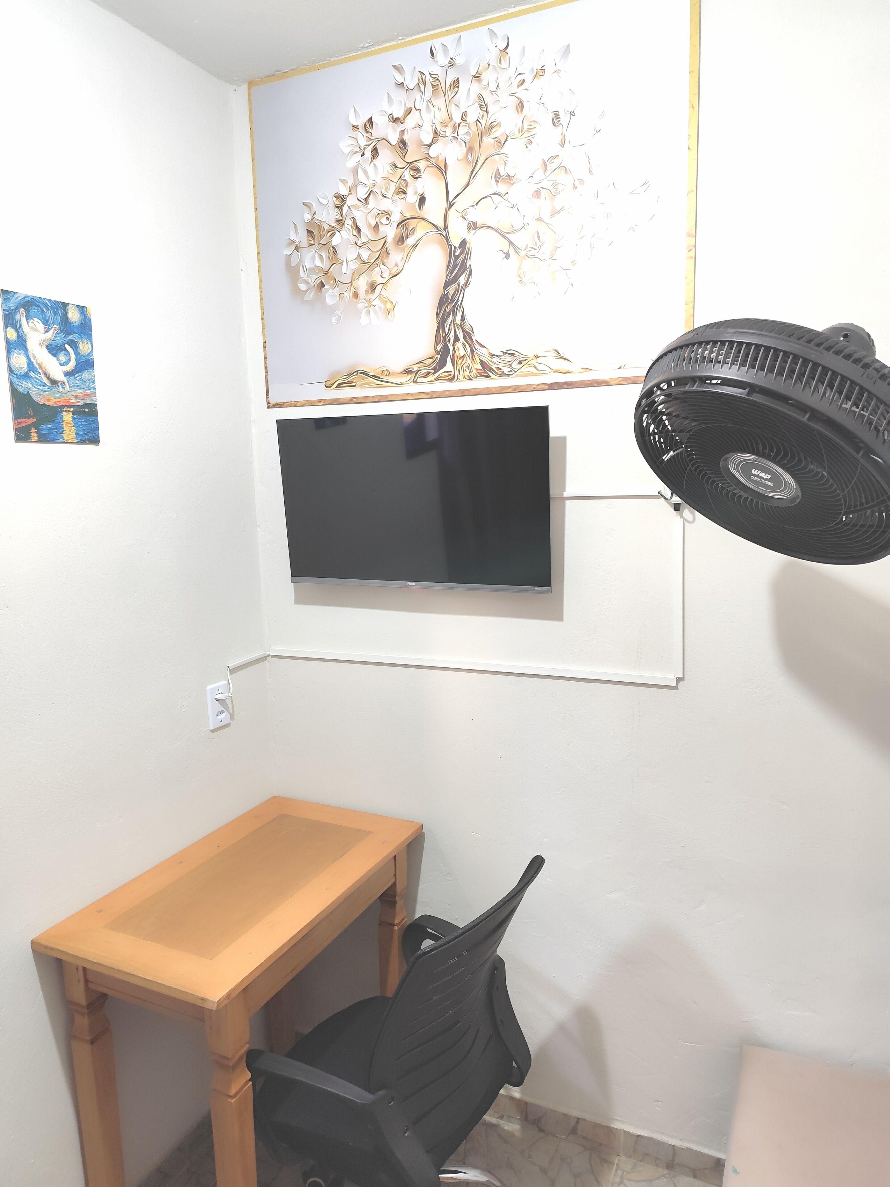A room with a wooden desk, a black office chair, a mounted TV, and a standing fan.