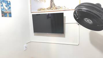 A room with a wooden desk, a black office chair, a mounted TV, and a standing fan.