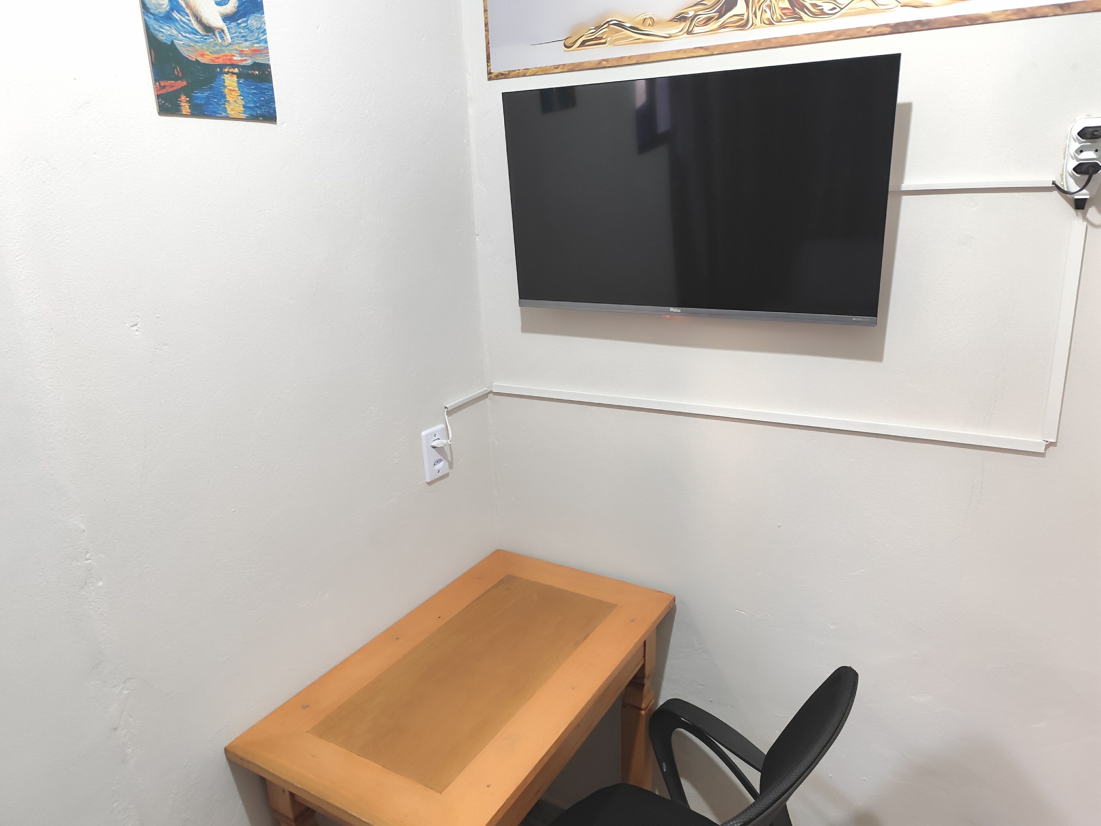 A small room with a wooden desk, a black chair, a wall-mounted TV, and a power outlet.