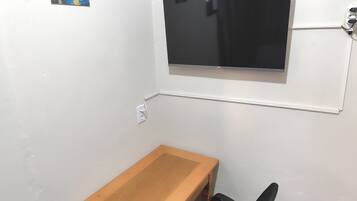 A small room with a wooden desk, a black chair, a wall-mounted TV, and a power outlet.