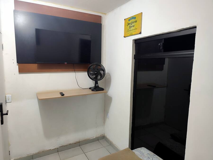 Una habitación con una televisión de pantalla plana montada en la pared, un ventilador de pie, una estantería con un control remoto y una puerta con un cartel encima.