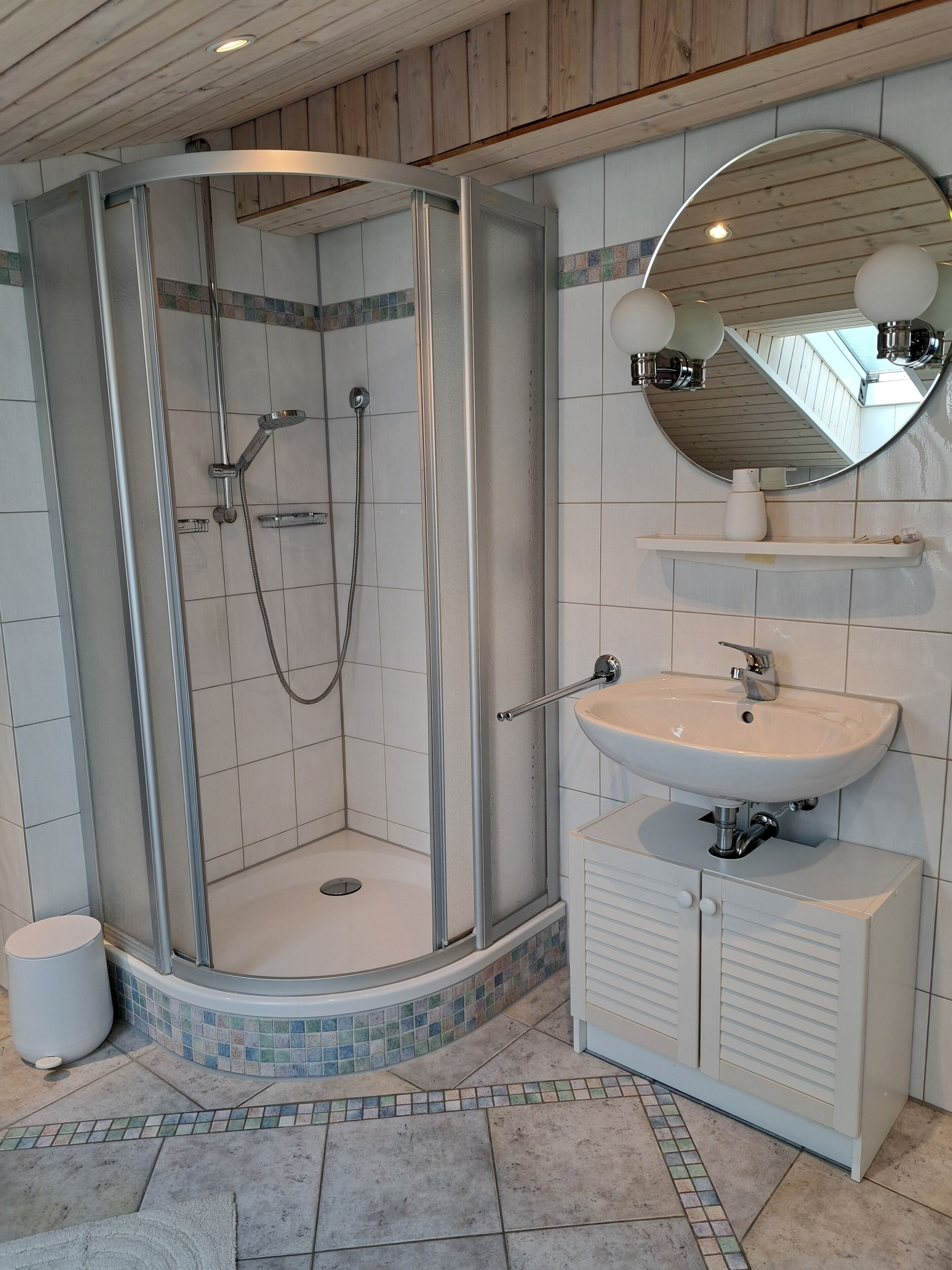 Eine Dusche mit Glastür, ein Waschbecken mit Spiegel darüber und ein gefliester Boden.